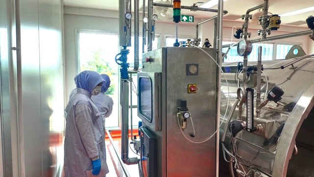 A person in a white robe and blue hood standing in a factory

Description automatically generated